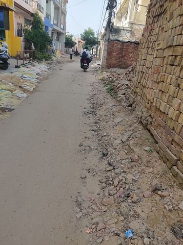 A person on a motorbike drives down a dusty road lined with rubble and brick walls, passing pedestrians and buildings. (Captioned by AI)
