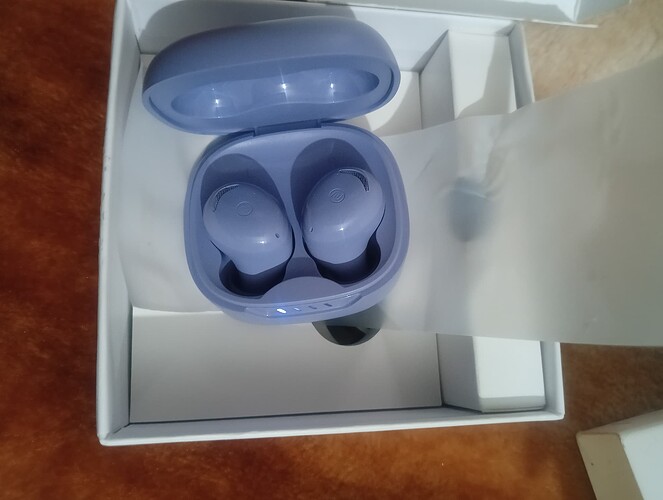 A pair of lavender wireless earbuds sits in their open charging case inside a white box. (Captioned by AI)