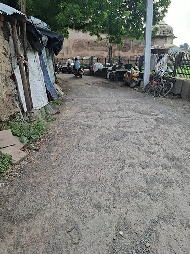 A narrow, gravel road runs between dilapidated shacks and a high wall, with parked motorcycles and bicycles lining the roadside. (Captioned by AI)