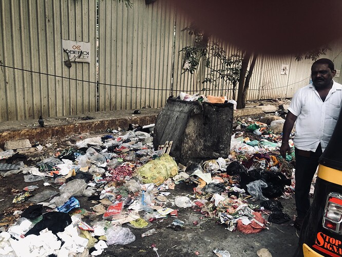 A man stands near an overflowing trash bin amidst a large pile of refuse scattered on a city street. (Captioned by AI)