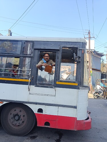 A bus driver leans out of the window of his bus, holding something in his hand, while passengers sit inside. (Captioned by AI)