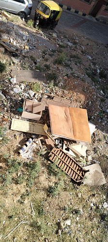 A pile of discarded furniture and debris sits amidst scattered trash on a patch of scrubby ground next to a street. (Captioned by AI)
